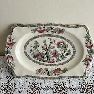 Puede incluir: Bandeja rectangular crema con un diseño floral y arbóreo en el centro. El diseño ovalado central está enmarcado por un borde negro y gris. Los diseños florales en rosa, azul y verde adornan los bordes.