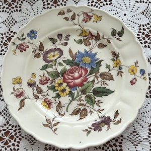 May include: A round, cream-coloured ceramic plate with a scalloped edge, featuring a central botanical illustration of a large pink rose, a blue flower, and various smaller yellow, purple, and red blossoms with green leaves and brown stems.