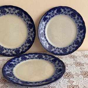 May include: Three antique ceramic plates with a cream-colored center and a blue floral pattern around the rim. The plates are arranged on a white lace tablecloth. The plates are a vintage style.