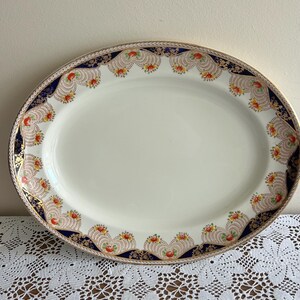 Peut inclure: Plat ovale de service avec un centre crème et une bordure décorative. La bordure présente une bande bleu marine avec des accents dorés et des motifs floraux rouges, orange et jaunes. Une pièce vintage.