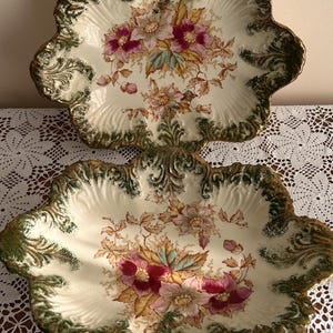 Puede incluir: Dos fuentes antiguas con borde festoneado. Los platos tienen un fondo crema con un diseño floral en rosa, amarillo y azul. Los bordes están adornados con detalles dorados y verdes. Se colocan sobre un mantel de encaje blanco.