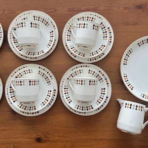 Op de afbeelding: Een vintage witte theeset met een geometrisch patroon in zwart, bruin en oranje. De set bevat kopjes, schotels, een groot bord en een melkkan. De randen van het servies hebben een gouden rand.