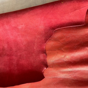 Può includere: Due pezzi di pelle rossa, uno piegato sull'altro. Il pezzo superiore è un camoscio liscio, mentre il pezzo inferiore ha una superficie più strutturata e granulosa.