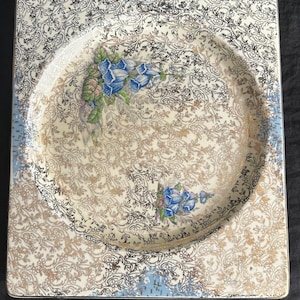 Pode incluir: Prato quadrado vintage com um design floral. O prato apresenta um poço central com detalhes florais azuis e um padrão de filigrana dourada e creme. A borda tem uma borda azul clara.