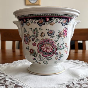 Pode incluir: Um vaso de cerâmica branca com borda alargada e duas alças laterais. O exterior apresenta um padrão floral colorido em tons de rosa, azul e verde, acentuado por uma faixa escura perto do topo e um fino acabamento dourado na base e na borda.