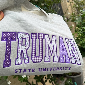 May include: A gray sweatshirt with the word "TRUMAN" in large, purple letters. The letters have different patterns, including polka dots, floral, and gingham. The text "STATE UNIVERSITY" is below the main text.