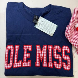 May include: Navy blue t-shirt with the words "OLE MISS" in red and white gingham letters. A tag with washing instructions is attached. A red and white gingham fabric is visible on the right side.