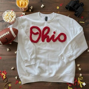 Peut inclure: Sweat-shirt blanc à col rond avec le mot "Ohio" en lettres rouges texturées. L'image comprend un ballon de football américain, du pop-corn, des chips et des jumelles, suggérant un thème de jour de match.