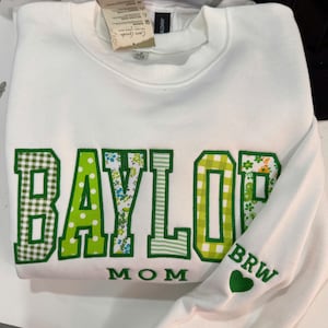 May include: A white crewneck sweatshirt featuring the word BAYLOR in large, green-bordered block letters, each filled with different green and white patterns including gingham, polka dots, and floral prints. The word MOM is below, with BRW and a heart on the sleeve.