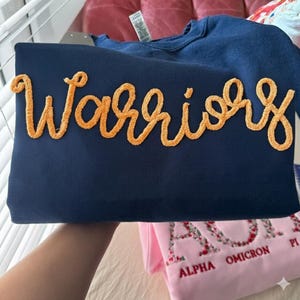 May include: A navy blue crewneck sweatshirt featuring the word Warriors in a textured, orange, cursive-style chenille patch appliqué across the chest. The garment has a standard long-sleeve design with a visible white brand tag at the neckline.