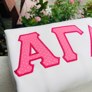 May include: White sweatshirt with large pink letters. The letters are decorated with white polka dots and outlined in a darker pink. The letters appear to spell out a Greek letter combination.