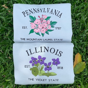 May include: Two light blue t-shirts with embroidered state flower designs. The top shirt features a pink mountain laurel flower with the text "PENNSYLVANIA EST. 1787 THE MOUNTAIN LAUREL STATE". The bottom shirt features a purple violet flower with the text "ILLINOIS EST. 1818 THE VIOLET STATE".