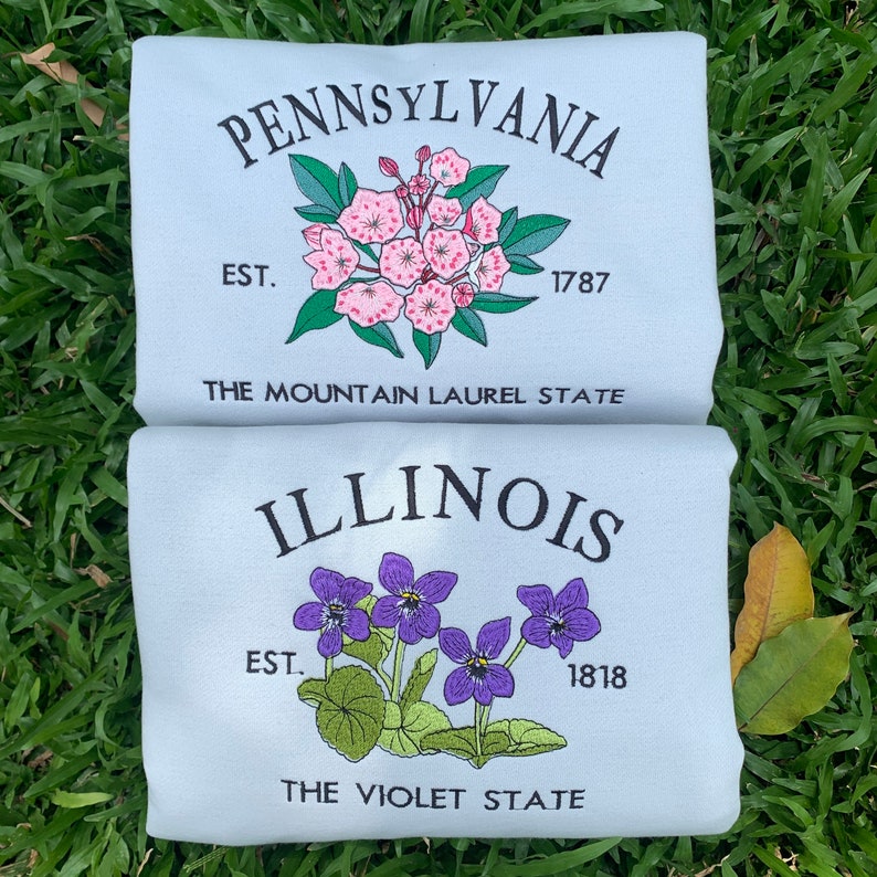 May include: Two light blue t-shirts with embroidered state flower designs. The top shirt features a pink mountain laurel flower with the text "PENNSYLVANIA EST. 1787 THE MOUNTAIN LAUREL STATE". The bottom shirt features a purple violet flower with the text "ILLINOIS EST. 1818 THE VIOLET STATE".