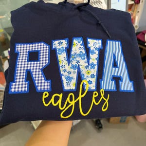 May include: A navy blue hooded sweatshirt featuring the letters RWA in large, fabric-appliqued block font. The R is blue and white gingham, the W is blue and white floral, and the A is blue and white striped. The word eagles is embroidered in yellow.