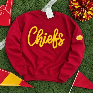 May include: A red crewneck sweatshirt featuring the word Chiefs in yellow cursive script across the chest. A small yellow and white football graphic is embroidered on the left sleeve. A white rectangular tag is attached to the neckline.