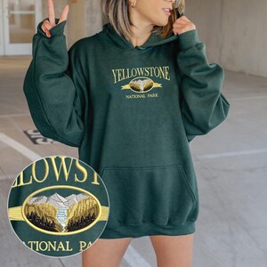 May include: Dark green hoodie with the text "YELLOWSTONE NATIONAL PARK" in gold lettering. The hoodie features a front pocket and a drawstring hood. A close-up shows an embroidered design of a mountain landscape.