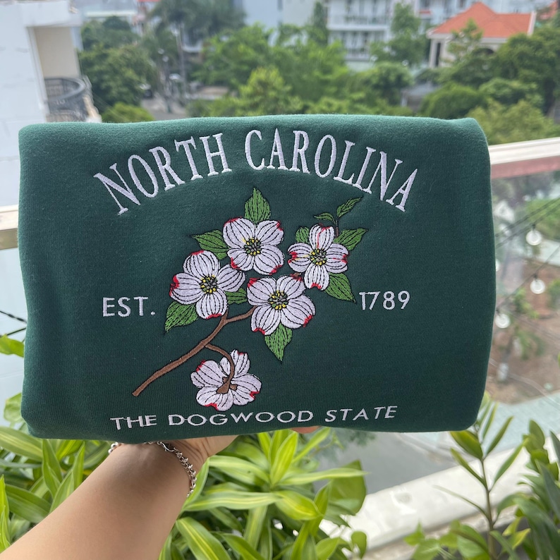 K&ouml;nnte beinhalten: Dunkelgr&uuml;nes Sweatshirt mit wei&szlig;em gesticktem Schriftzug "NORTH CAROLINA" und "THE DOGWOOD STATE". Das Design umfasst ein Blumenmotiv aus wei&szlig;en Blumen mit roten und gr&uuml;nen Akzenten und das Jahr "1789".