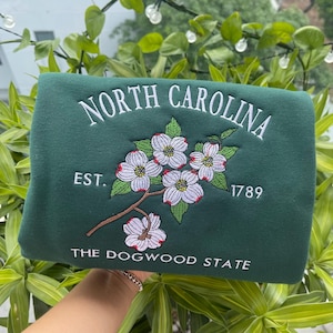 K&ouml;nnte beinhalten: Dunkelgr&uuml;nes Sweatshirt mit wei&szlig; gesticktem Text "NORTH CAROLINA" &uuml;ber einem Blumendesign. Darunter stehen die Worte "EST. 1789" und "THE DOGWOOD STATE".