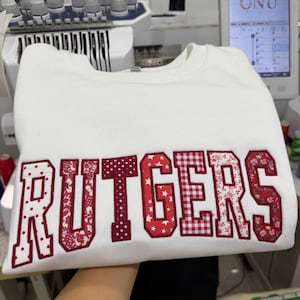 May include: A white sweatshirt with the word "RUTGERS" embroidered across the front in a mix of patterned fabrics, including polka dots, floral, and gingham. The sweatshirt is displayed in a workshop setting.