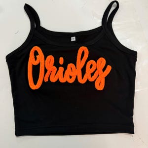 Op de afbeelding: Een zwarte mouwloze crop top met het woord Orioles in feloranje, getextureerde chenille letters op de borst. Het kledingstuk heeft een ronde halslijn, dunne schouderbandjes en een maat M label zichtbaar bij de binnenkant van de kraag.