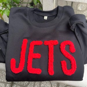 May include: Black sweatshirt featuring the word "JETS" in bold, red, textured letters. The sweatshirt has a crew neck and long sleeves. The image is a close-up of the front of the garment.