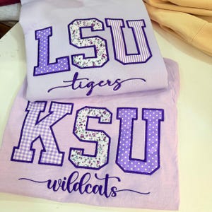 Pode incluir: Duas camisolas de cor roxo claro com letras de estilo universitário. A camisola de cima exibe LSU com padrões florais e às riscas, e a de baixo exibe KSU com padrões axadrezados, florais e às bolinhas, ambas com texto em cursivo.