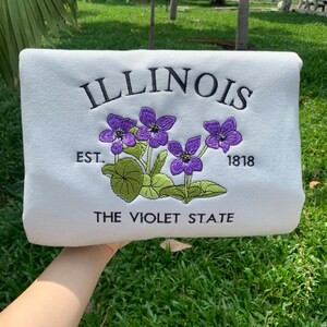 May include: White sweatshirt with embroidered text "ILLINOIS" and "THE VIOLET STATE" with a purple flower design and the text "EST. 1818".