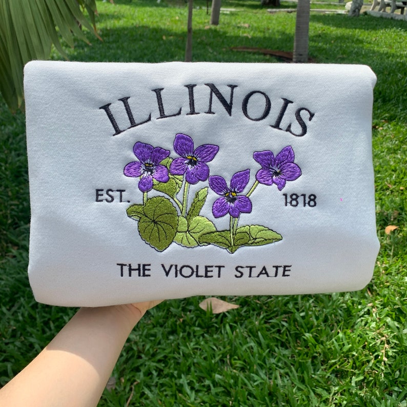 May include: White sweatshirt with embroidered text "ILLINOIS" and "THE VIOLET STATE" with a purple flower design and the text "EST. 1818".