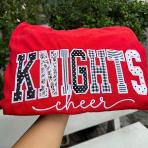 May include: A bright red sweatshirt featuring the word KNIGHTS in large, block-style letters with varied black and white patterns including stars, florals, stripes, and polka dots, with the word cheer written below in white cursive script.