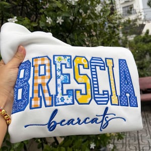 Könnte beinhalten: Ein weißes Sweatshirt mit dem Wort BRESCIA in großen, blockartigen Stickbuchstaben, die jeweils ein einzigartiges Muster wie Vichy-Karo, Blumen und Streifen in Blau, Gelb und Orange aufweisen. Darunter steht das Wort bearcats in blauer Schreibschrift.
