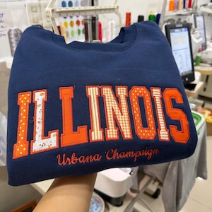 May include: A navy blue crewneck sweatshirt featuring the word ILLINOIS in large, block-style applique letters. The letters alternate between solid orange, orange and white gingham, and white with orange floral patterns. Urbana Champaign is embroidered below.