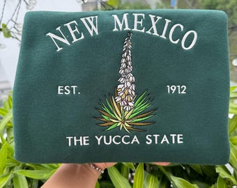 May include: Green sweatshirt with white embroidered text that reads "New Mexico" and "The Yucca State". The sweatshirt also features a white embroidered yucca plant with the text "Est. 1912" above it.
