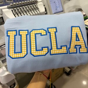May include: A light blue sweatshirt with the embroidered letters "UCLA" in a yellow and white gingham pattern, outlined in blue. The sweatshirt is held up in front of an embroidery machine.