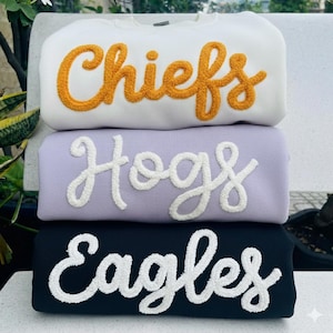 May include: Three sweatshirts stacked on top of each other. The top sweatshirt is white with the word "Chiefs" in orange script. The middle sweatshirt is lavender with "Hogs" in white script. The bottom sweatshirt is navy blue with "Eagles" in white script.