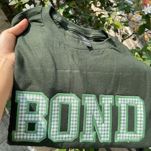 Könnte beinhalten: Dunkelgrünes T-Shirt mit dem Wort „BOND“ in großen, grün-weiß karierten Buchstaben auf der Vorderseite. Das T-Shirt ist gefaltet, und der Kragen ist sichtbar. Der Stoff scheint Baumwolle zu sein.