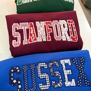 Könnte beinhalten: Drei gefaltete Sweatshirts in Dunkelgrün, Kastanienbraun und Blau. Das grüne Sweatshirt zeigt das Wort BINGHAMTON in grün-weiß gemusterten Buchstaben. Das kastanienbraune zeigt STANFORD in rot-weißen Mustern. Das blaue zeigt SUSSEX in marineblau-weißen Mustern.