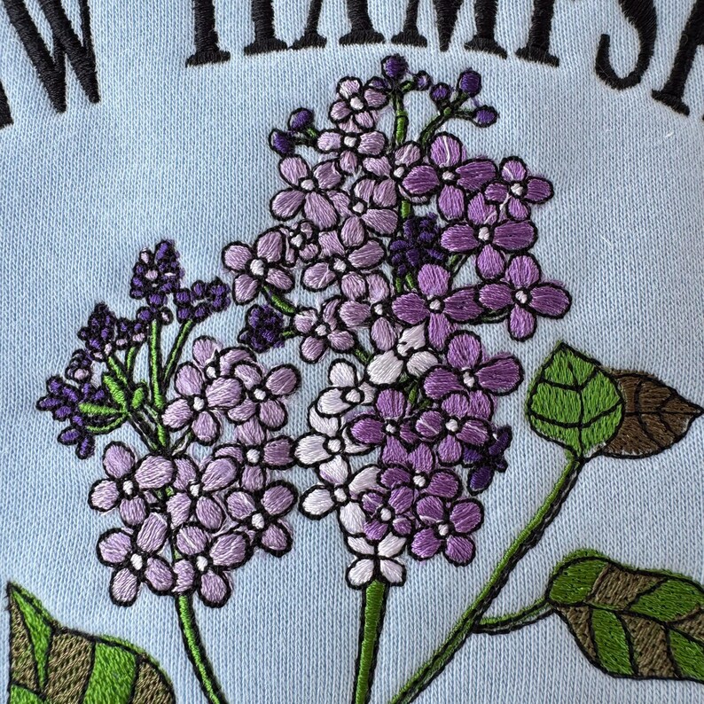 May include: Embroidered lilac flowers in shades of purple and white on a light blue background. The flowers are arranged in a cluster and have a delicate, feminine look.