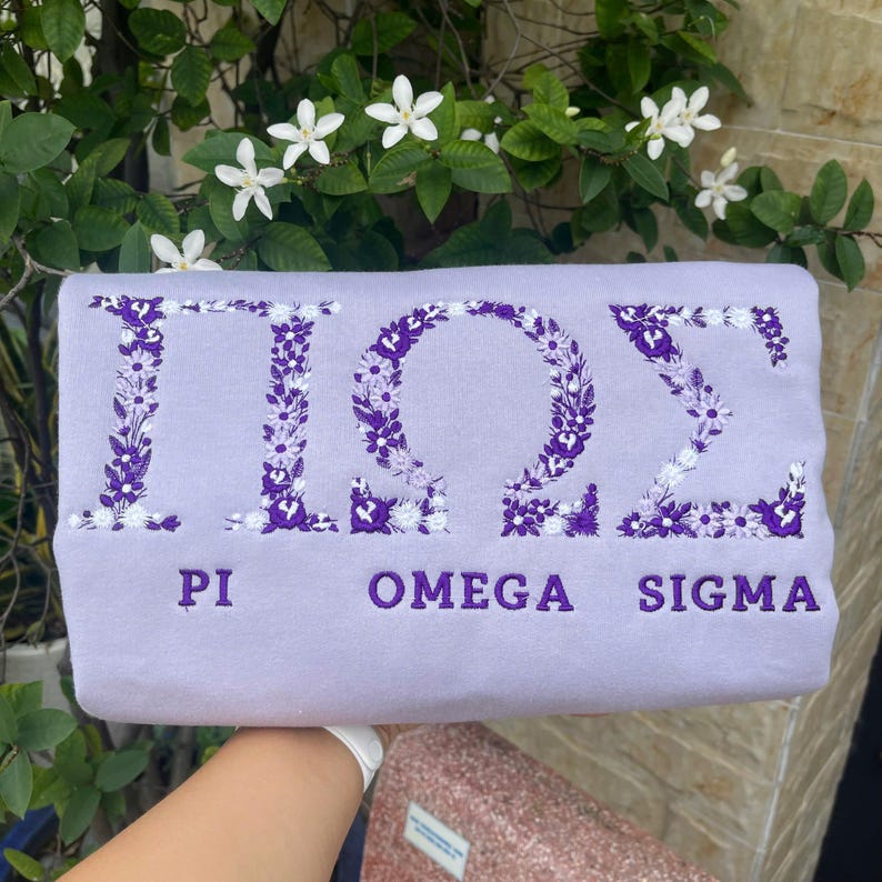Pu&ograve; includere: Una felpa lilla con lettere greche ricamate e dettagli floreali. Le lettere sono riempite di fiori viola e bianchi. Sotto le lettere ci sono le parole "PI", "OMEGA" e "SIGMA" in viola.