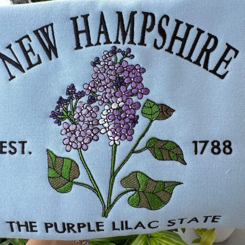 May include: A light blue sweatshirt with embroidered text that reads "NEW HAMPSHIRE" in black, with a purple lilac flower design in the center. The text "EST. 1788" is embroidered in black below the flower, and the text "THE PURPLE LILAC STATE" is embroidered in black below the year.