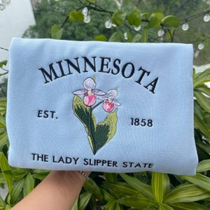 May include: Light blue sweatshirt with embroidered text "MINNESOTA EST. 1858 THE LADY SLIPPER STATE" and a floral design of two pink and white lady slipper orchids.