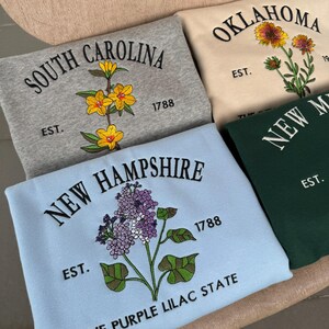 May include: Four sweatshirts with embroidered state names and flowers. The sweatshirts are in different colors: gray, beige, blue, and green. The flowers are yellow, purple, and orange.