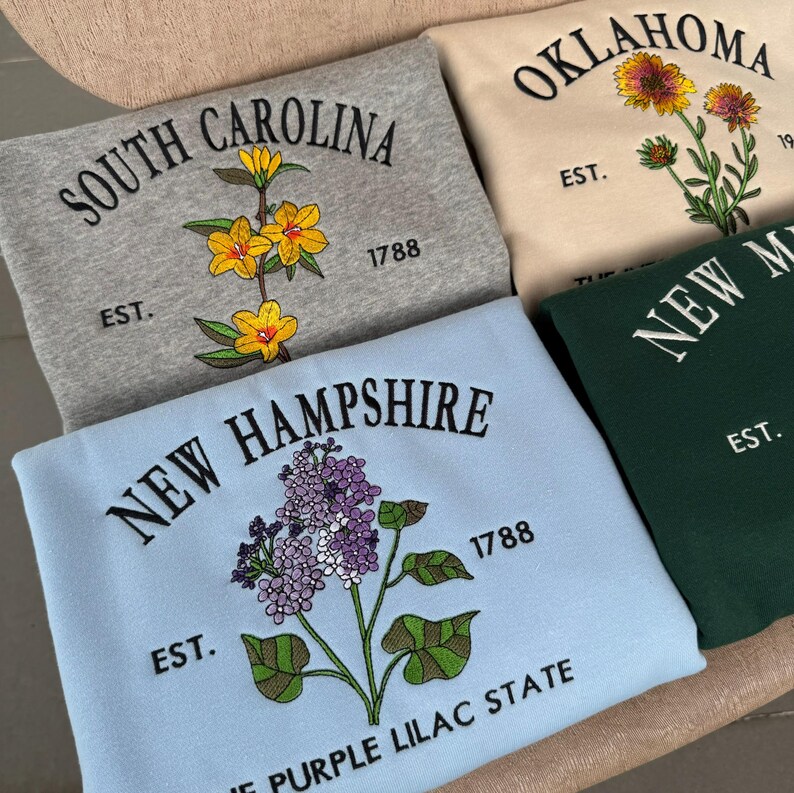 May include: Four sweatshirts with embroidered state names and flowers. The sweatshirts are in different colors: gray, beige, blue, and green. The flowers are yellow, purple, and orange.