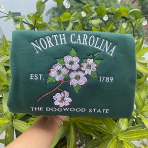 K&ouml;nnte beinhalten: Ein dunkelgr&uuml;nes Sweatshirt mit wei&szlig; gesticktem Schriftzug "NORTH CAROLINA". Es zeigt einen Zweig des Blumen-Hartriegels mit wei&szlig;en und rosa Bl&uuml;ten, den Text "EST. 1789" und "THE DOGWOOD STATE".
