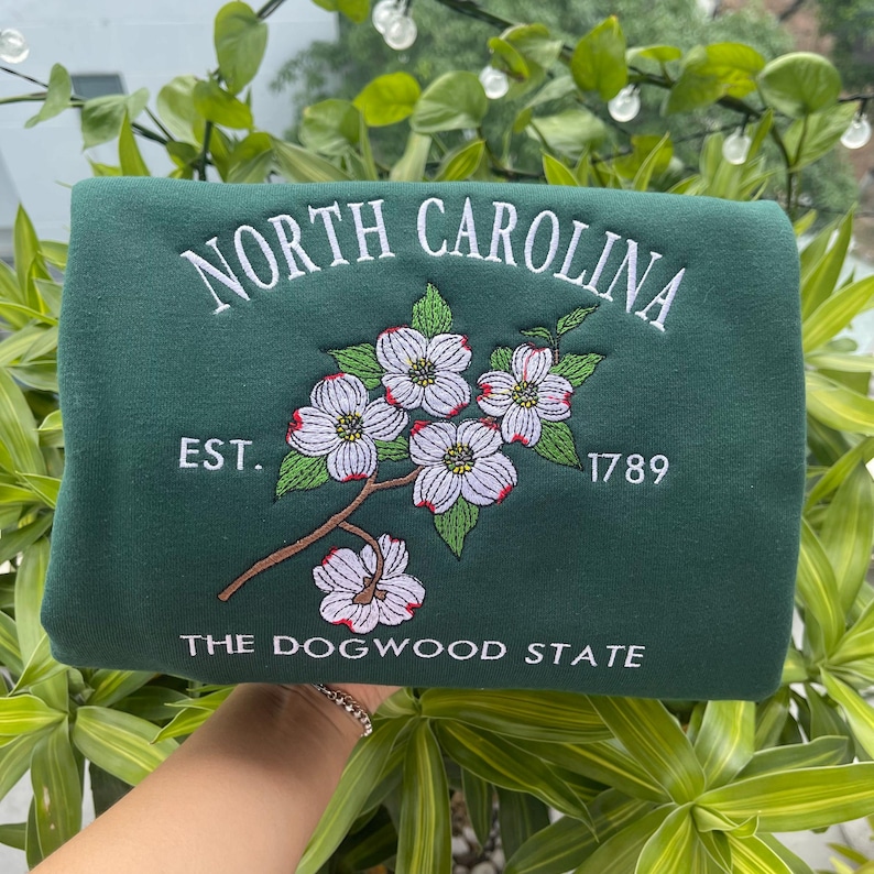 K&ouml;nnte beinhalten: Ein dunkelgr&uuml;nes Sweatshirt mit wei&szlig; gesticktem Schriftzug "NORTH CAROLINA". Es zeigt einen Zweig des Blumen-Hartriegels mit wei&szlig;en und rosa Bl&uuml;ten, den Text "EST. 1789" und "THE DOGWOOD STATE".