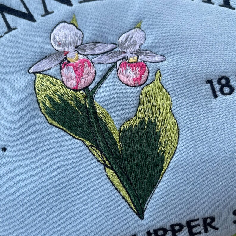 May include: Embroidered design of two pink lady slipper orchids with green leaves on a light blue background. The text "18" is visible in the top right corner.
