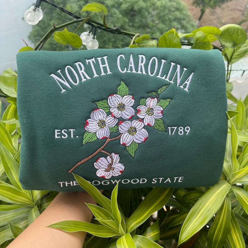 K&ouml;nnte beinhalten: Ein dunkelgr&uuml;nes Sweatshirt mit wei&szlig; gesticktem Text &bdquo;NORTH CAROLINA&ldquo; &uuml;ber einem floralen Design. Darunter stehen die Worte &bdquo;EST. 1789&ldquo; und &bdquo;THE DOGWOOD STATE&ldquo;. Das florale Design zeigt wei&szlig;e Bl&uuml;ten mit roten Zentren.