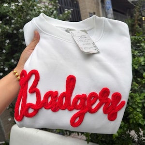 Pode incluir: Uma camisola branca de gola redonda com a palavra Badgers em letras grandes, vermelhas, texturizadas e felpudas de estilo chenille na frente. Uma pequena etiqueta branca presa à gola exibe o texto Crafted & Decor Stitched with love made for you.