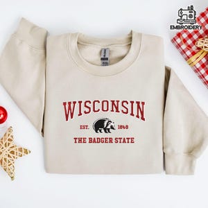 May include: A beige sweatshirt with the word "WISCONSIN" in maroon block letters. Below the word is a black and white badger graphic and the text "EST. 1848 THE BADGER STATE". The sweatshirt is folded on a white surface.