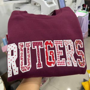 May include: A maroon sweatshirt with the word "RUTGERS" across the front. The letters are white with red polka dots, floral, and star patterns. The sweatshirt is made of a soft material.