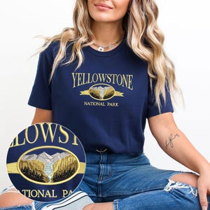 May include: Navy blue t-shirt with the embroidered text "YELLOWSTONE NATIONAL PARK" in gold. The design features a mountain and river scene within a gold oval. The shirt is a short-sleeved crew neck.